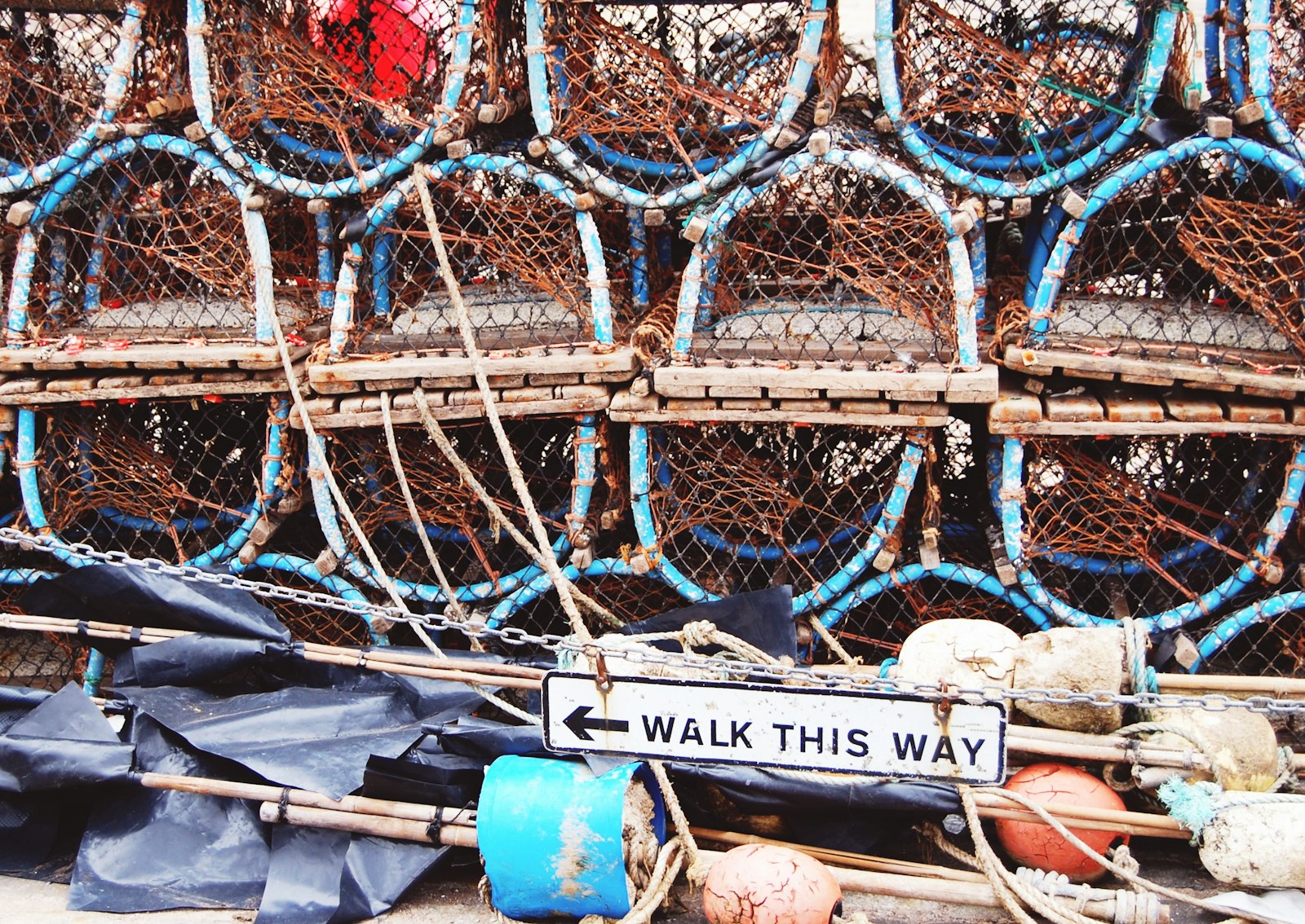 A wall of stacked lobster traps on a dock, with a sign in front reading 'Walk This Way' and an arrow pointing along them